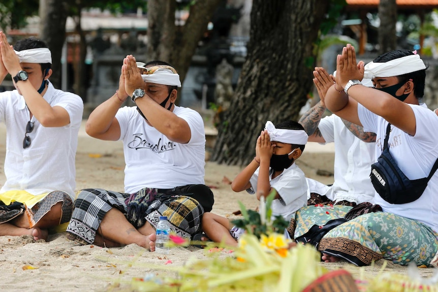 Four men and a boy sit cross legged on a beach with their eyes closed and hands pressed together in front of their faces.