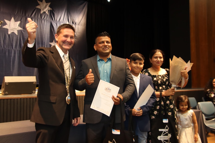 A smiling man in a suit with a thumb up standing next to a man, a young boy, a woman and a young girl holding certificates