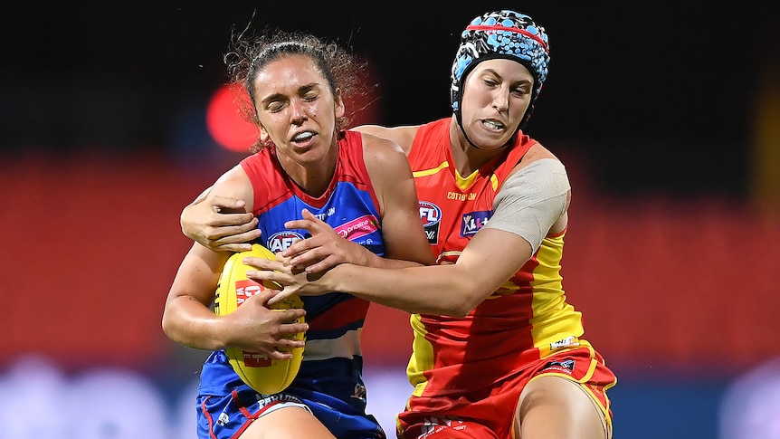 A Western Bulldogs AFLW player holds the ball as she is tackled by a Gold coast Suns opponent.