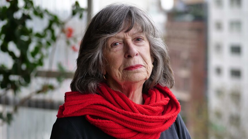 A portrait shot of an elderly woman wearing a red scarf. She has a serious expression on her face.