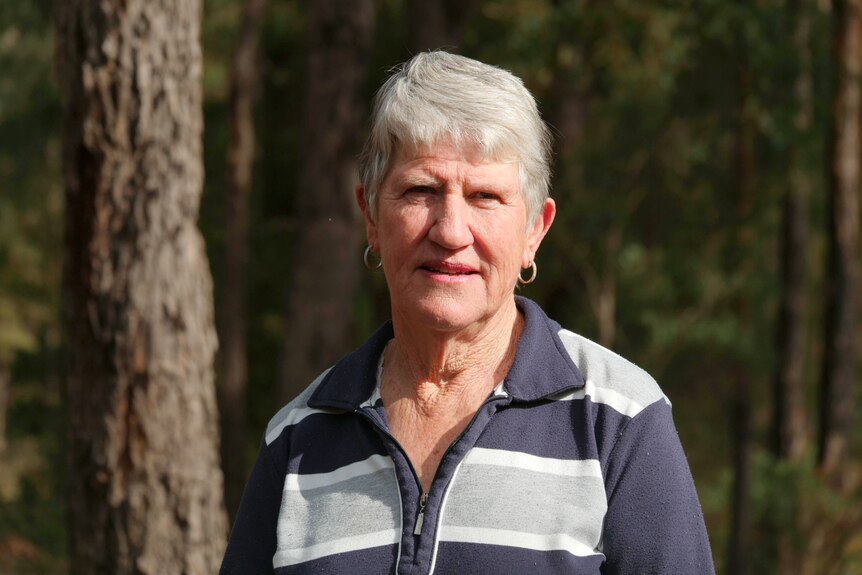 An older woman with short grey hair stands in a forested area.