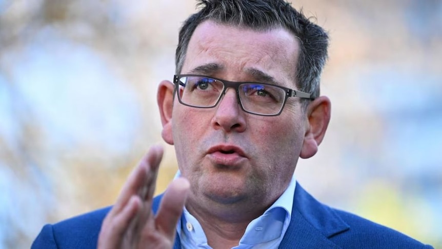 Victorian premier Daniel Andrews wears a navy suit in front of a blurred background. His hand is raised and he is mid speech. 