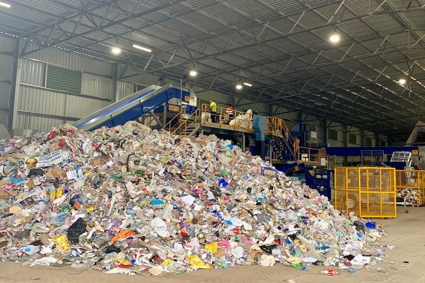 A pile of recyclable materials sits in front of a processing machine in a large shed.