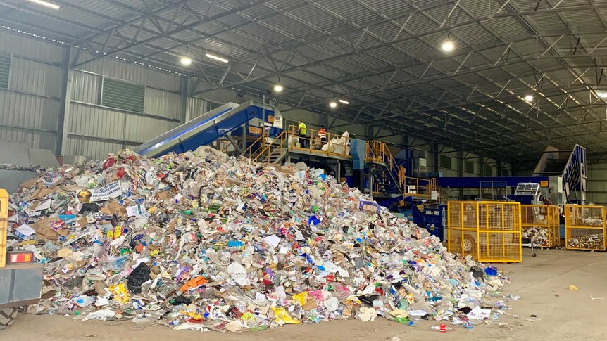 A pile of recyclable materials sits in front of a processing machine in a large shed.