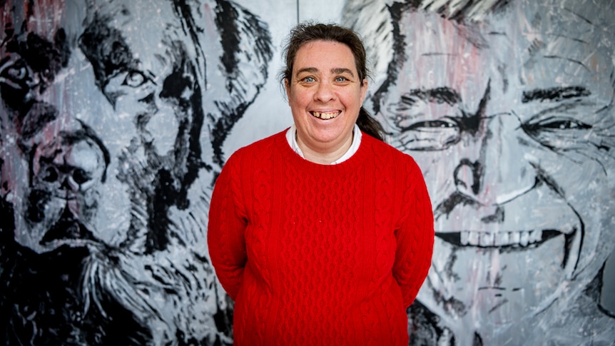 Fiona McKenzie stands in a red jumper in front of a black and white mural of a dog and a man, and she smiles at the camera.