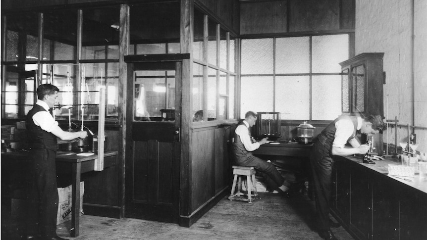 Three men in vests and shirts lean over lab equipment in a wood panelled room