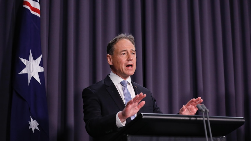 A man in a suit stands in front of a lectern speaking, a blue curtain is behind him.