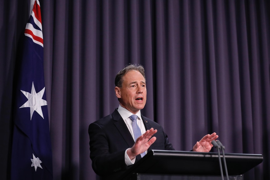 A man in a suit stands in front of a lectern speaking, a blue curtain is behind him.