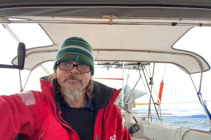 Self of man in red coat, glasses and green beanie at helm of boat at sea