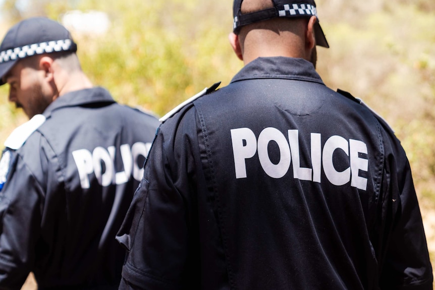 Two police recruits, wearing blue overalls with the word POLICE written in white on the back, with police hats in bushland.