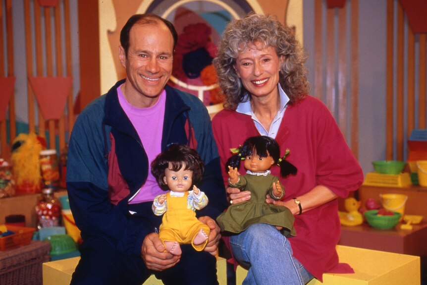 Play School presenters George and Benita sit next to each other.  They are smiling and both hold a child's doll.