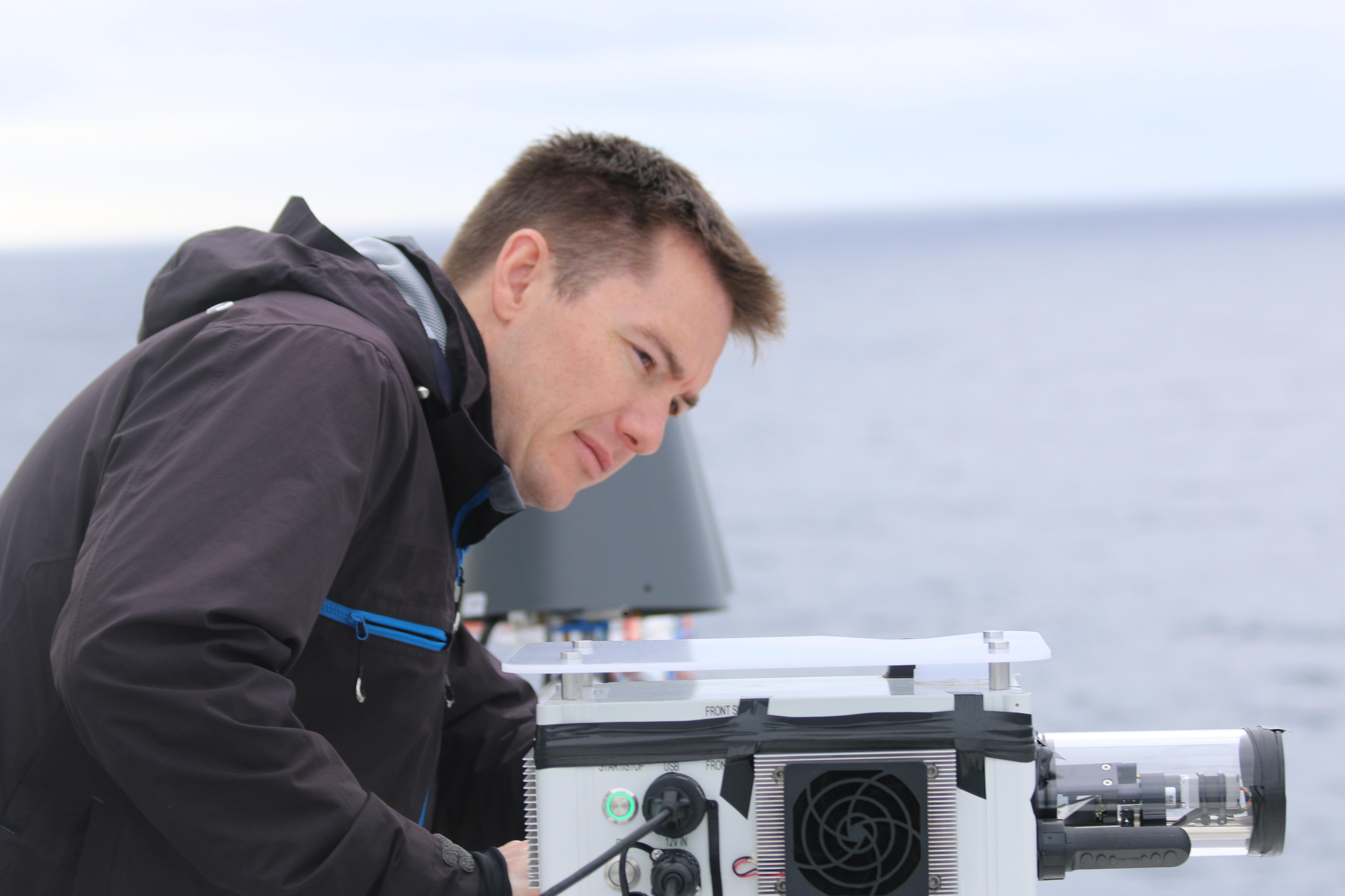 A man wearing a black jacket leans over a piece of machinery, there is water in the background.
