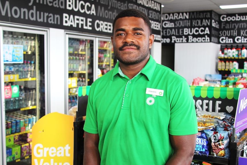 A man in a bright green polo shirt stares at the camera from inside a convenience store. 