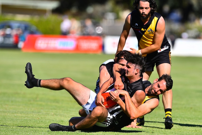 A footballer gets tackled by another footballer, they are wearing black white and yellow. Lying on a green grass oval.