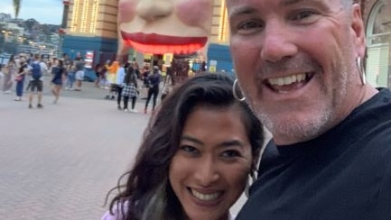 Smiling woman with long hair and man with two silver earing pose in a selfie photo outside Luna Park entrance in Sydney.