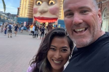 Smiling woman with long hair and man with two silver earing pose in a selfie photo outside Luna Park entrance in Sydney.