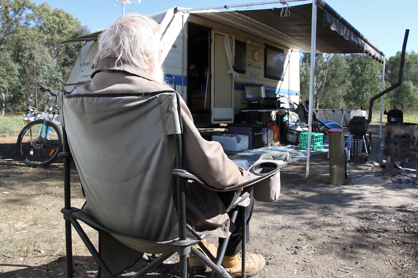 A man with grey hair sits outside a caravan, with a cooking fire