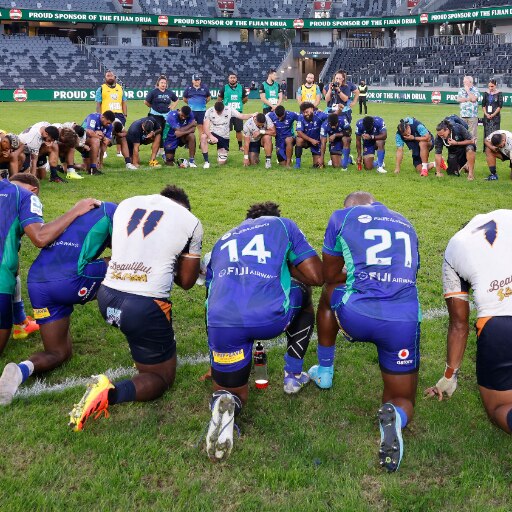 Men kneeling in prayer on a green rugby field in a circle wearing blue and white uniforms.