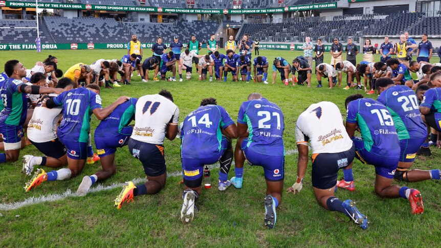 Men kneeling in prayer on a green rugby field in a circle wearing blue and white uniforms.