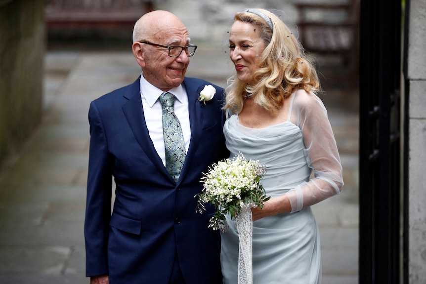 Rupert Murdoch in a blue suit and Jerry Hall in a wedding dress stand arm in arm outside a church