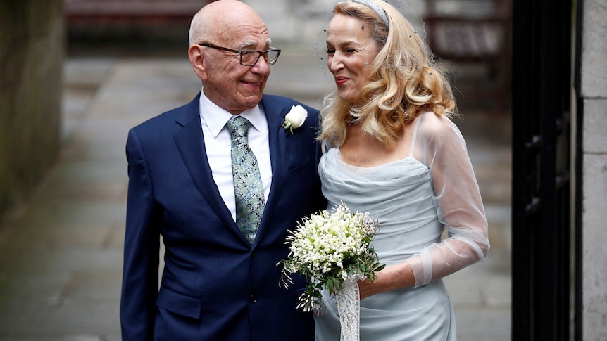 Rupert Murdoch in a blue suit and Jerry Hall in a wedding dress stand arm in arm outside a church