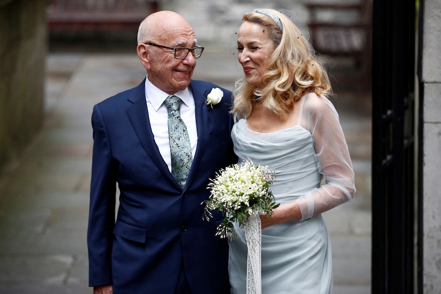 Rupert Murdoch con un traje azul y Jerry Hall con un vestido de novia están tomados del brazo afuera de una iglesia