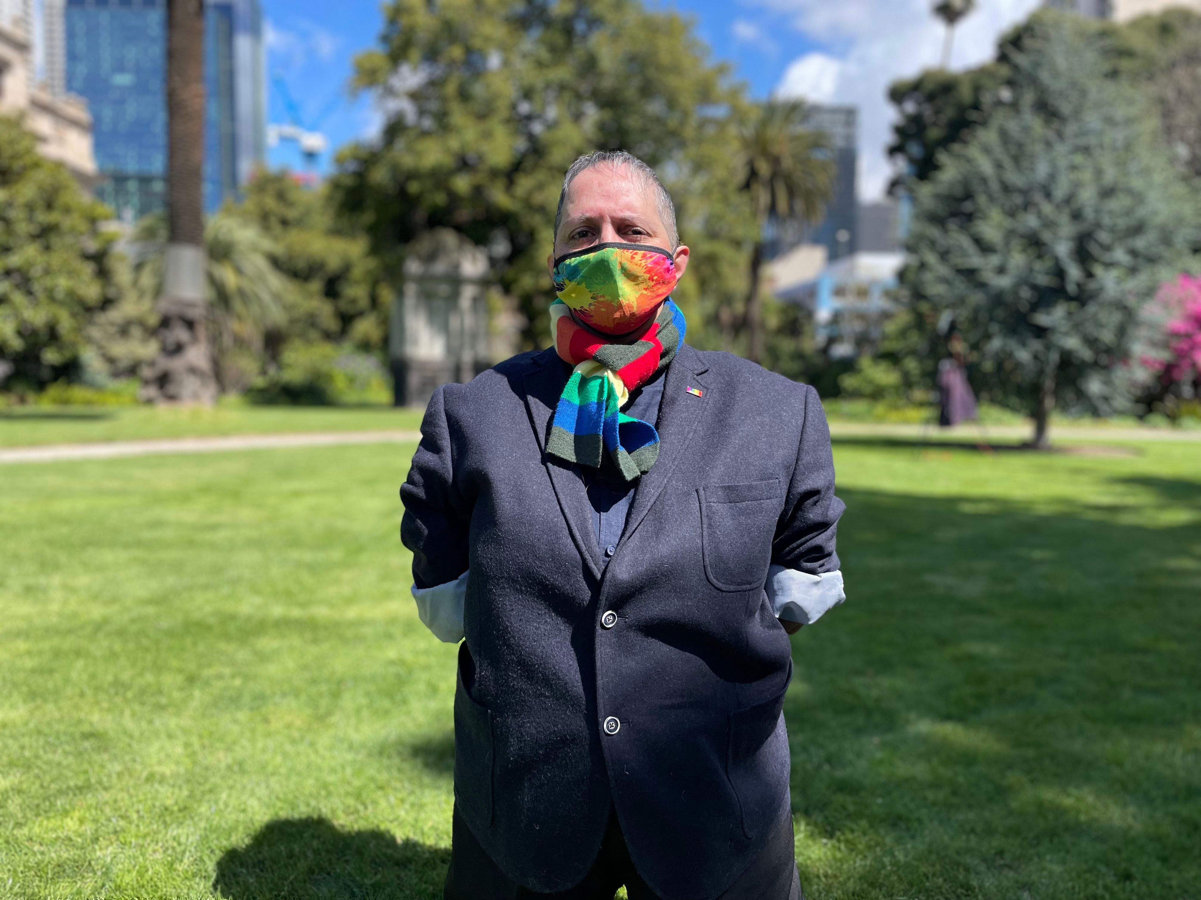 A woman with close-cropped hair stands with a rainbow-coloured mask on her face in a garden.