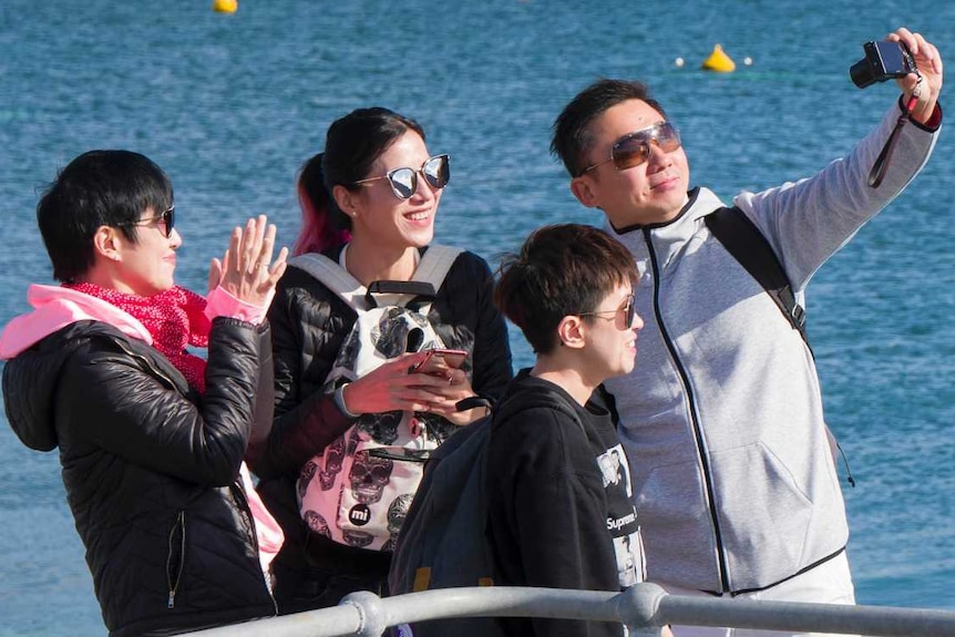 A group of four tourists stand on a jetty taking a selfie on Rottnest Island with the ocean behind them.