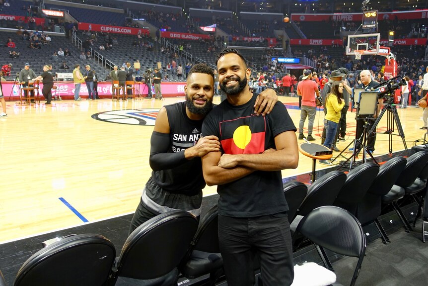 Patty and Luke are standing together and smiling in front of a basketball court. Luke is wearing an Aboriginal flag t-shirt.