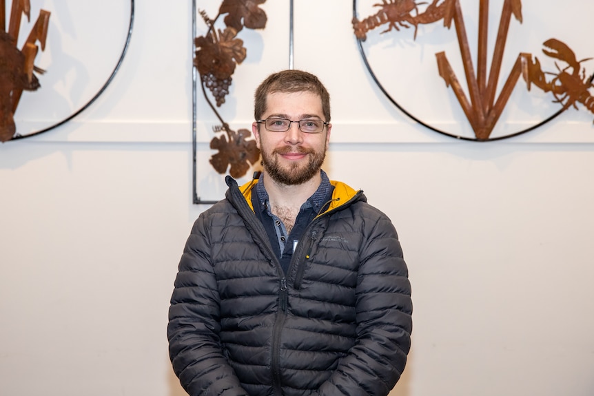 A man wearing color transition glasses and a puffer jacket is standing in front of a metal wall hanging.