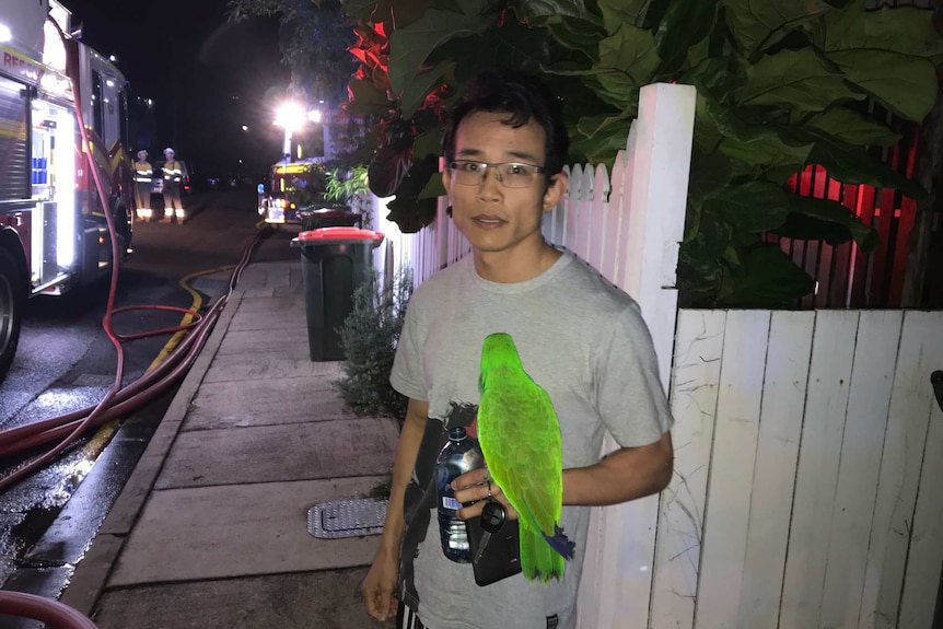 A man stands holding a water bottle, wallet and keys with a bright green parrot on his arm next to 2 fire trucks