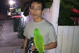 A man stands holding a water bottle, wallet and keys with a bright green parrot on his arm next to 2 fire trucks