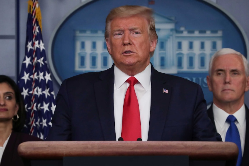 An elderly man in a dark suit and red tie speaks seriously in front of a man and woman and flag.