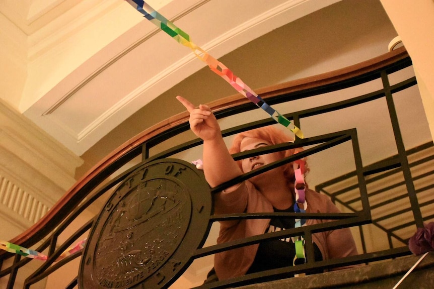 A young person with pink hair points through a railing that is covered in colourful paper chains.