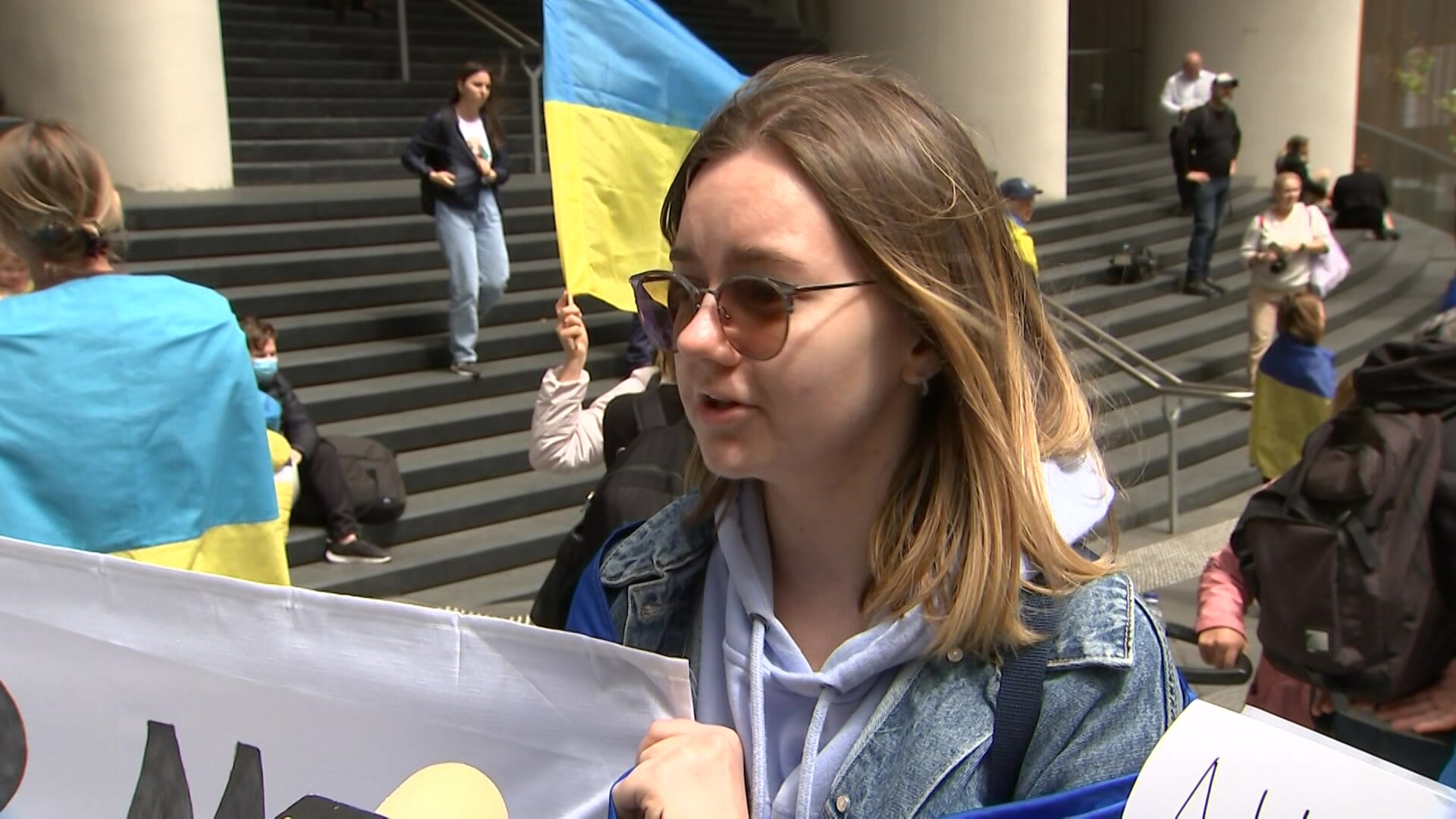 A woman in sunglasses and a denim jacket stands in front of a Ukrainian flag with a partially obscured placard.