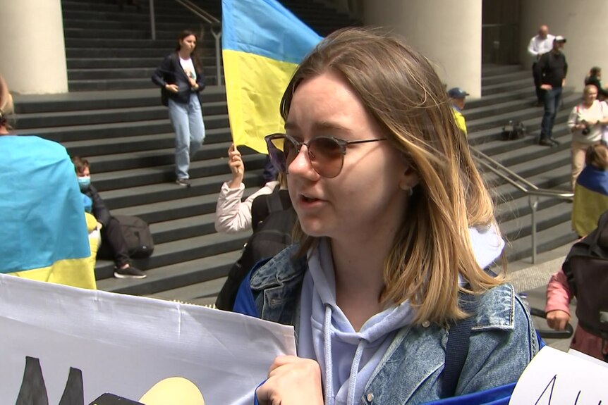 A woman in sunglasses and a denim jacket stands in front of a Ukrainian flag with a partially obscured placard.