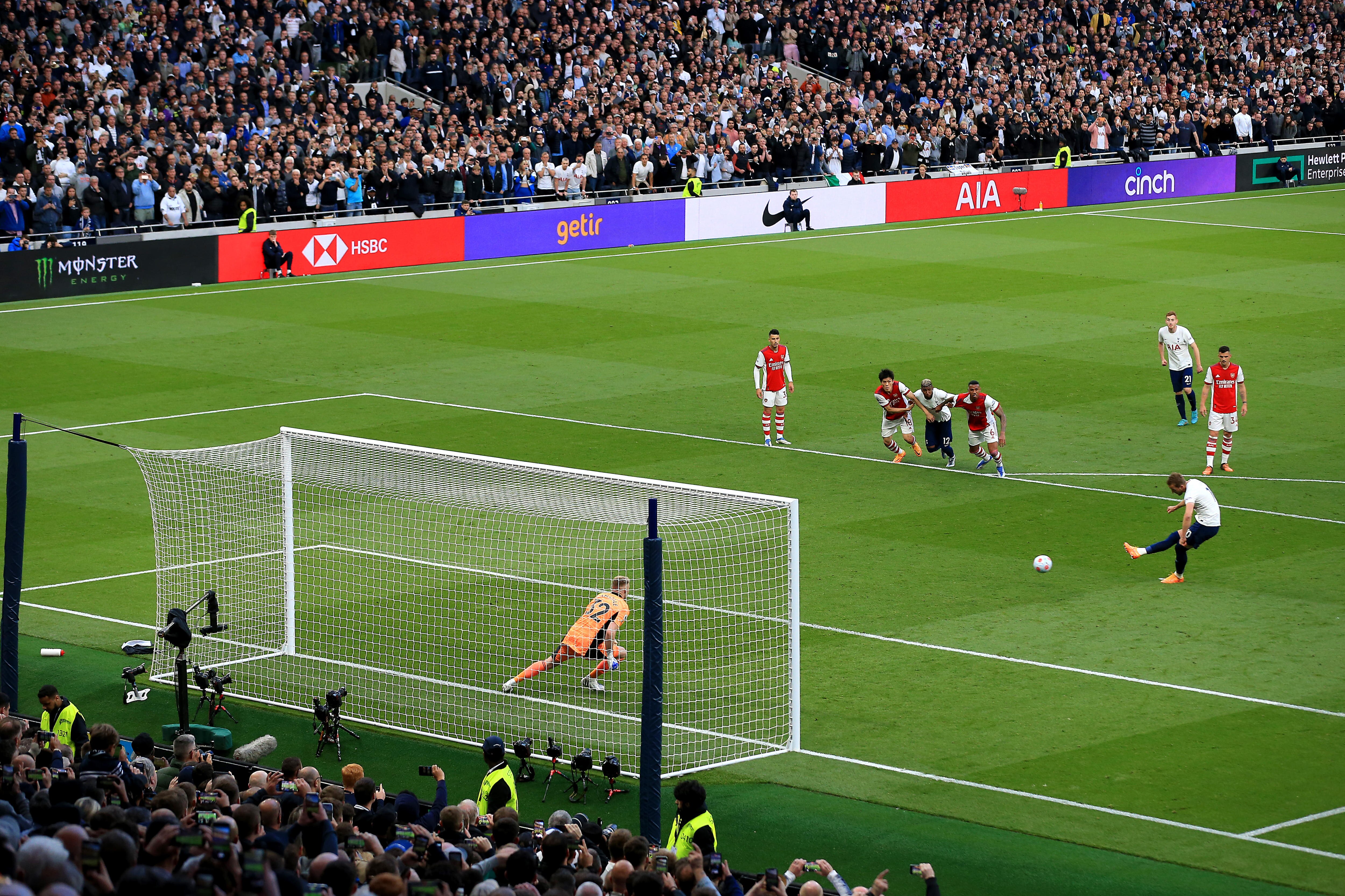 A male soccer player wearing white and blue kicks the ball from the penalty spot against a goalkeeper during a game