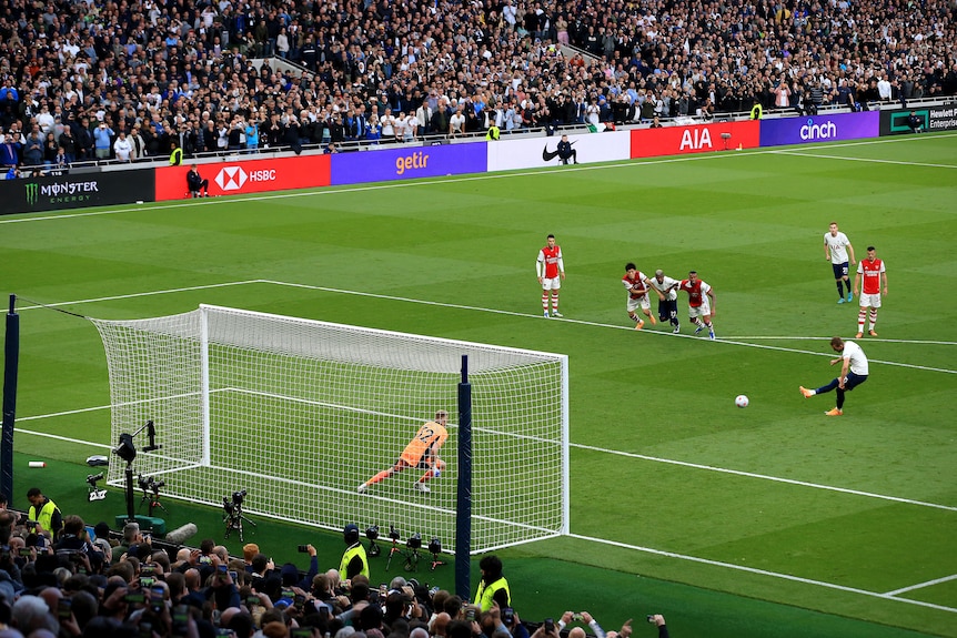 A male soccer player wearing white and blue kicks the ball from the penalty spot against a goalkeeper during a game