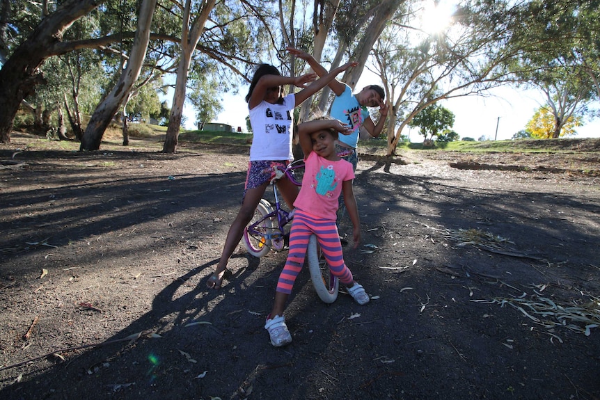 Three young kids, outdoors on bikes, dabbing.