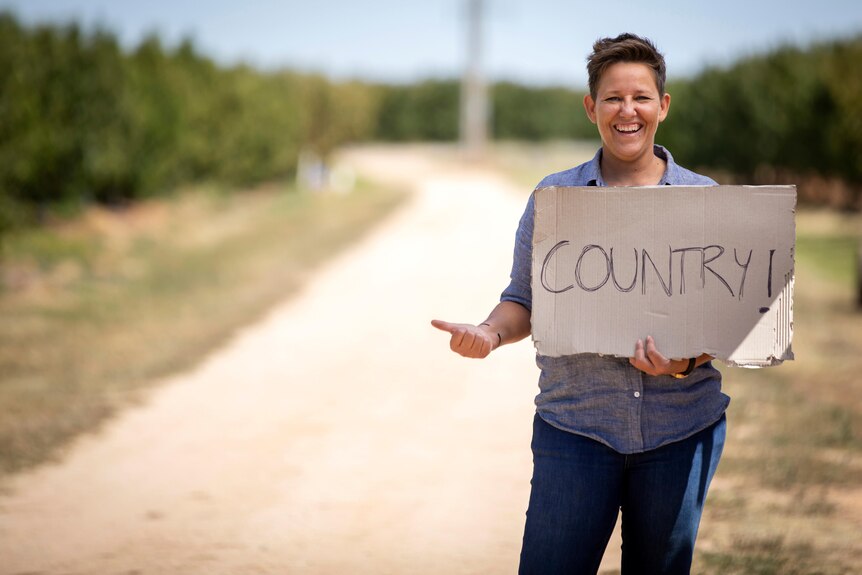 A woman wearing a blue shirt stands by a road with her thumb out, holding a sign that says 'country'