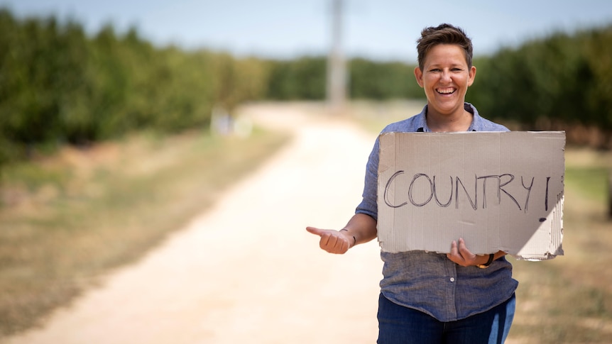 A woman wearing a blue shirt stands by a road with her thumb out, holding a sign that says 'country'