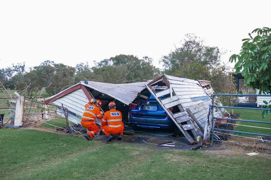 Two men in orange SES suits inspect a collapsed garage over a blue car.