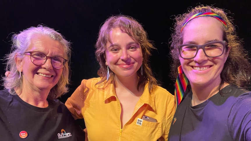 Three people are brightly lit against a black background which may be a theatre, each smiling at the camera.