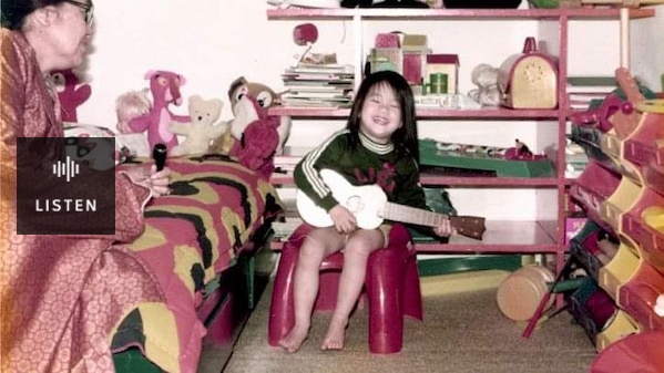 Lisa Leong as a child playing the guitar in her bedroom smiling. Has Audio.