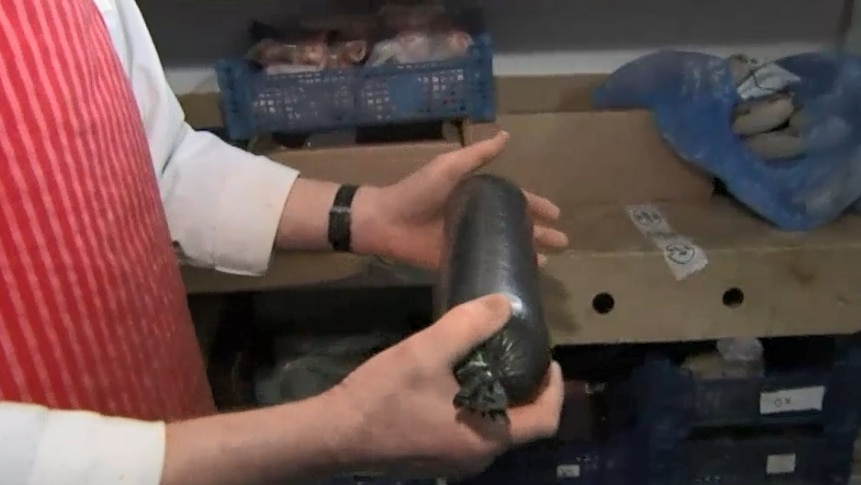 A man holds a large black sausage in a walk-in freezer