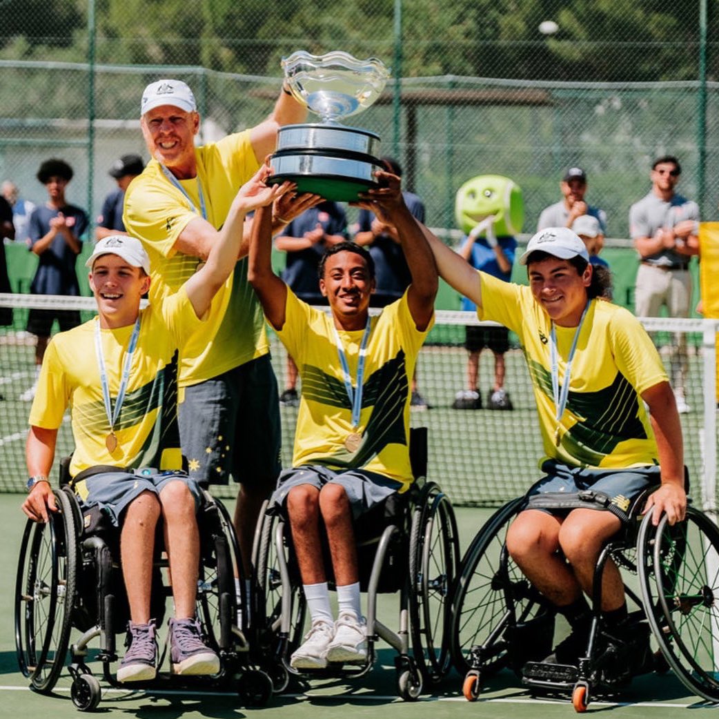 Three wheelchair tennis players raise their arms in celebration, holding a trophy along with their coach.