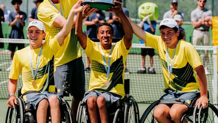 Three wheelchair tennis players raise their arms in celebration, holding a trophy along with their coach.