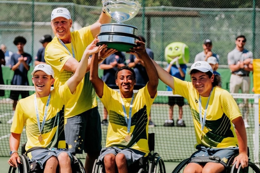 Three wheelchair tennis players raise their arms in celebration, holding a trophy along with their coach.