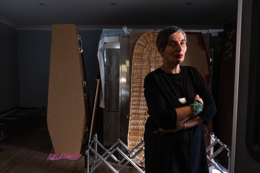 A woman wearing black stands with arms crossed in a room that has several types of coffins