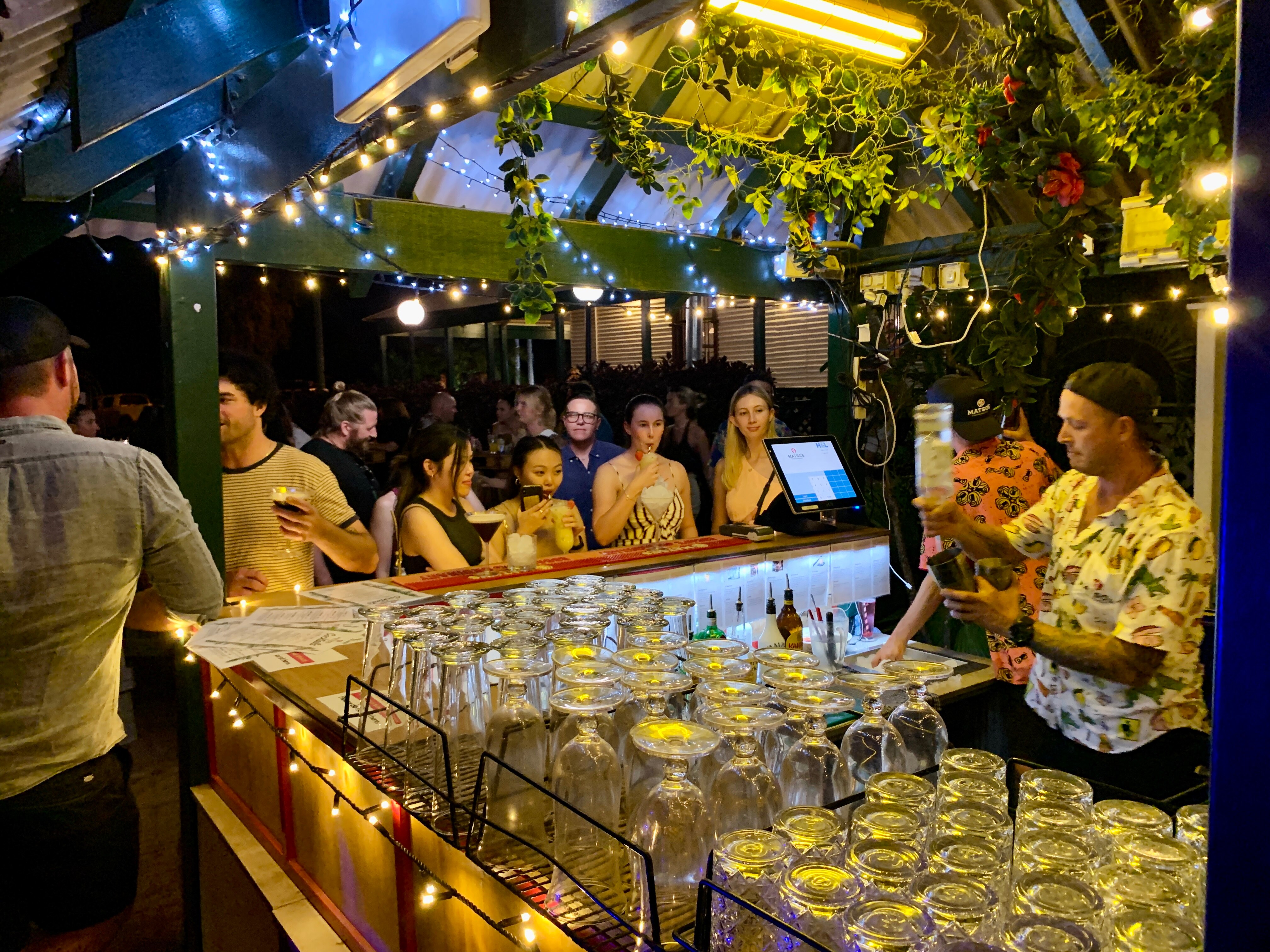 Young people line up at an outdoor bar festooned with pot plants and fairy lights